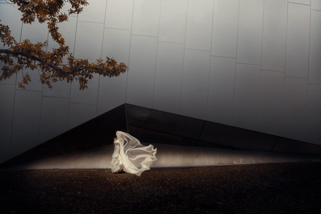 Bride looking over shoulder with veil blowing over her face framed within the triangle shape of the Melbourne museum building