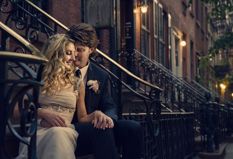 Happy moment of bride leaning into groom while sitting on stairs at dusk on the streets of New York on their wedding day