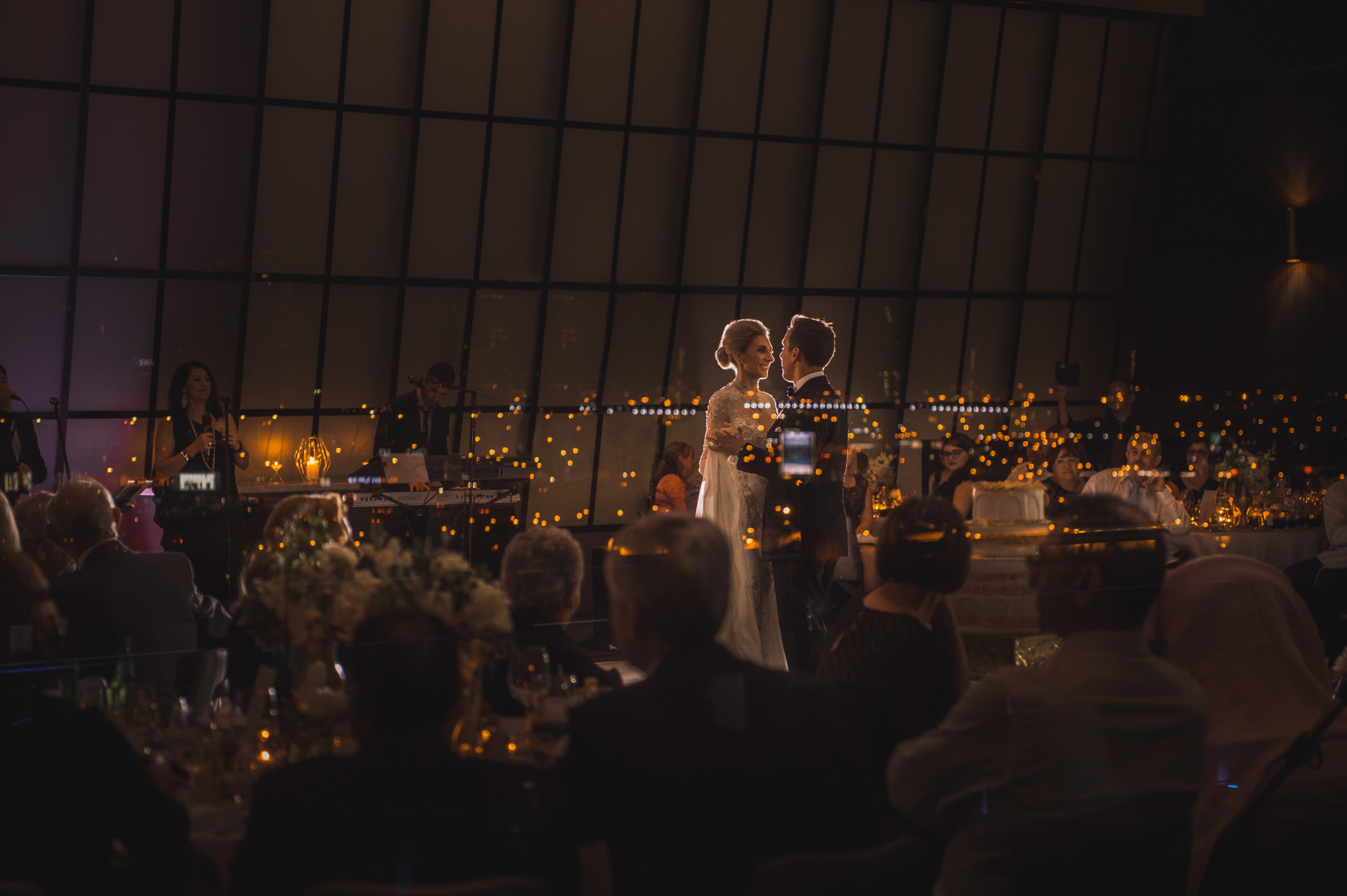 Candid photo of Wedding couples first dance seen through windows of Luminare reception with refection of the Melbourne city scape with guests