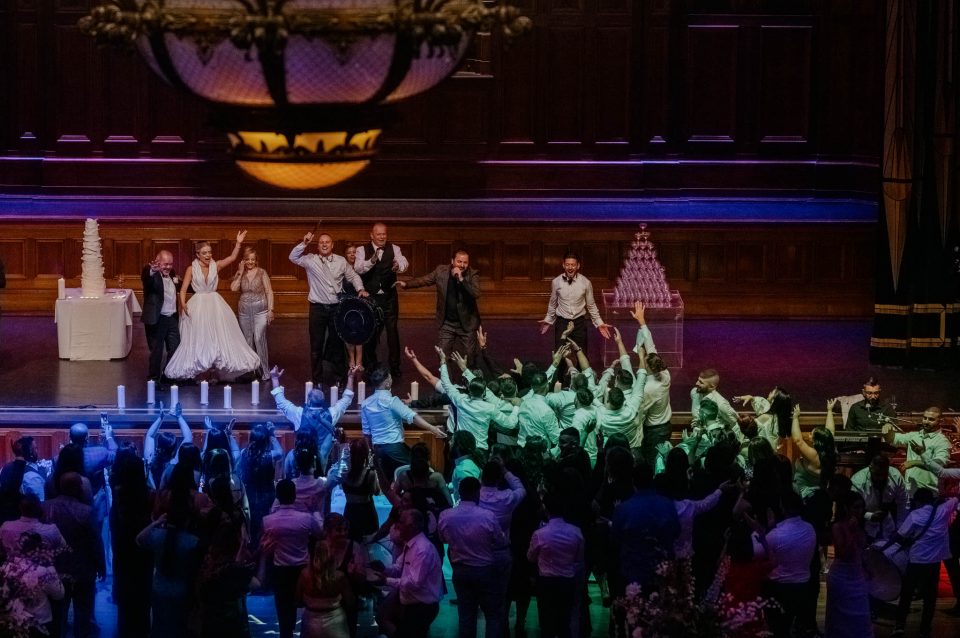 Bride and groom with their families on the stage dance to their guests on the floor below