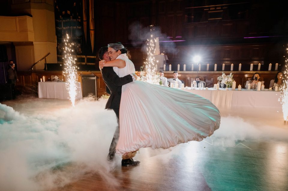 Groom lifts and swirls bride during the bridal waltz with dry ice effects surround them on the dance floor with indoor sparklers