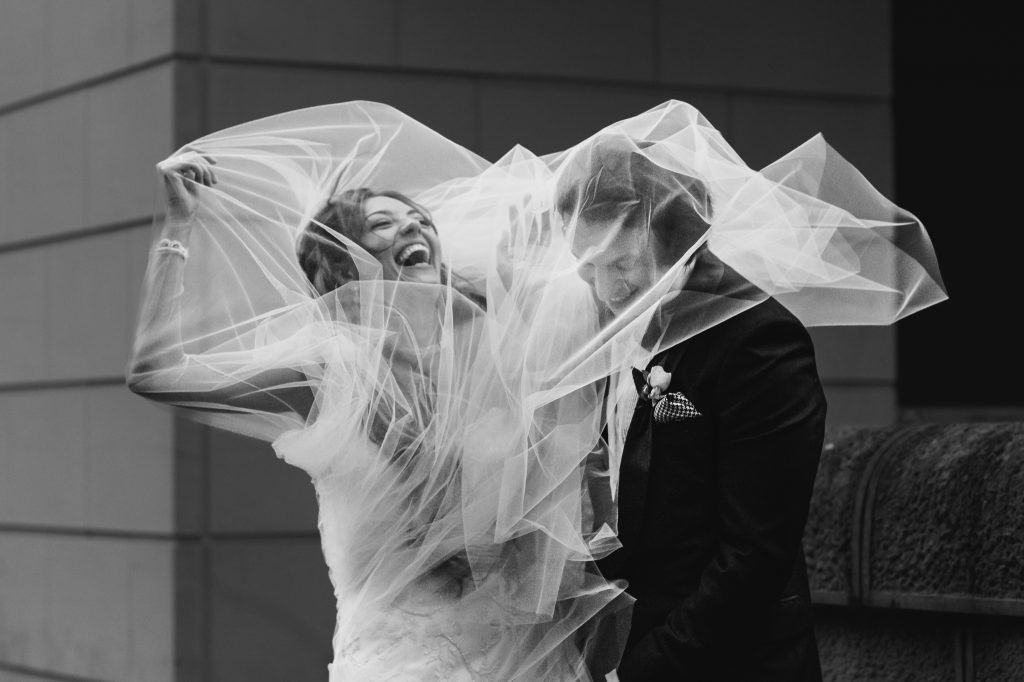 Black and White photograph of bride & groom laughing with both as they are stuck under veil