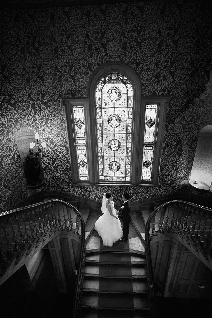 Bride & groom on the grand staircase at Labassa Mansion beneath an ornate stained-glass window in a timeless black and white portrait.