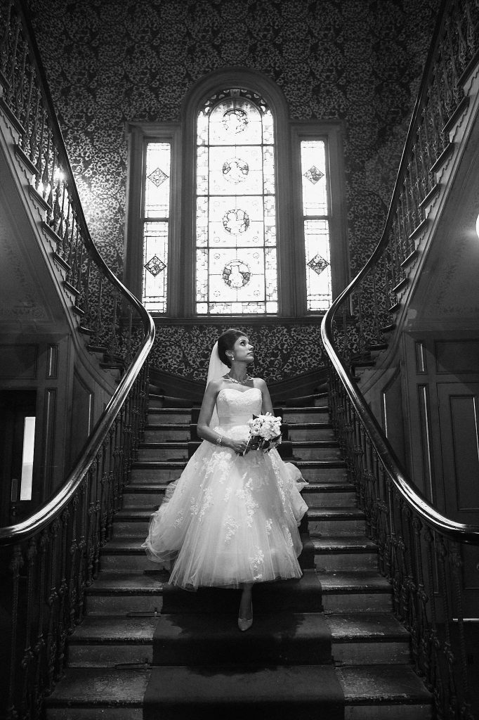 Bride descending the grand staircase at Labassa Mansion beneath a stained-glass window in a dramatic black and white portrait.